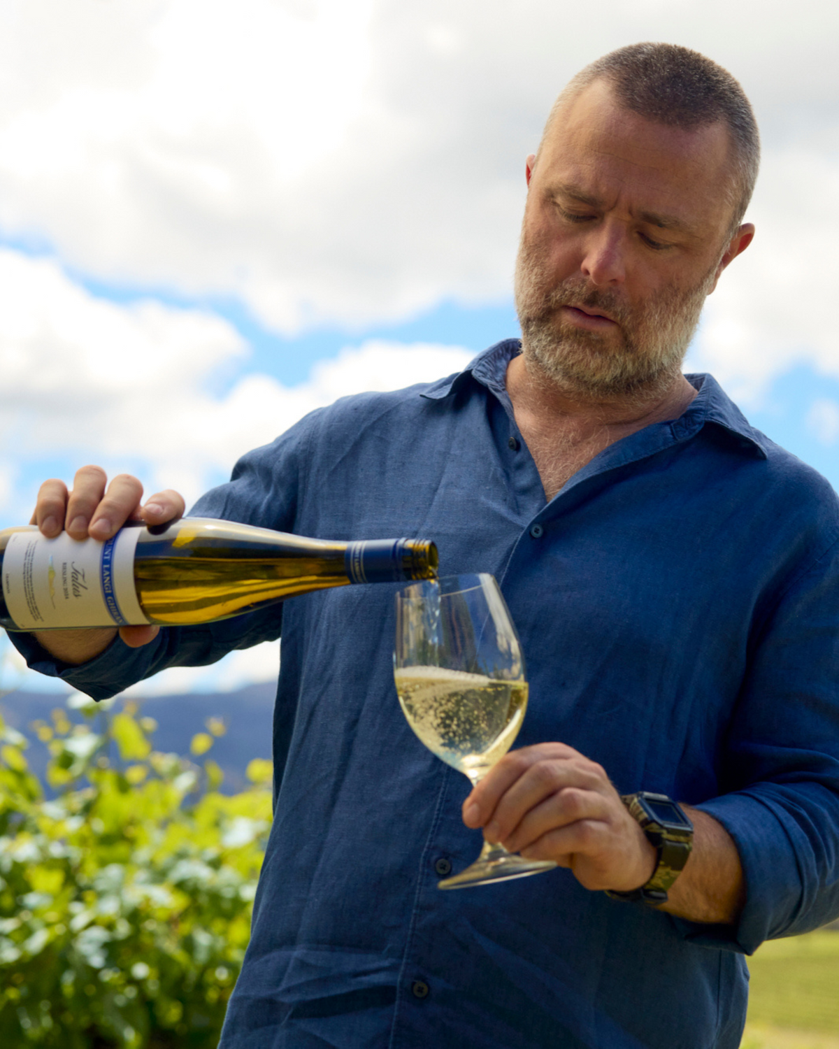 Man in a blue linen shirt pouring white wine into a glass in the vineyard