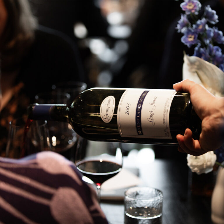 Hand tilting a bottle of aged Mount Langi Ghiran Shiraz over a table with a wine glass and flowers