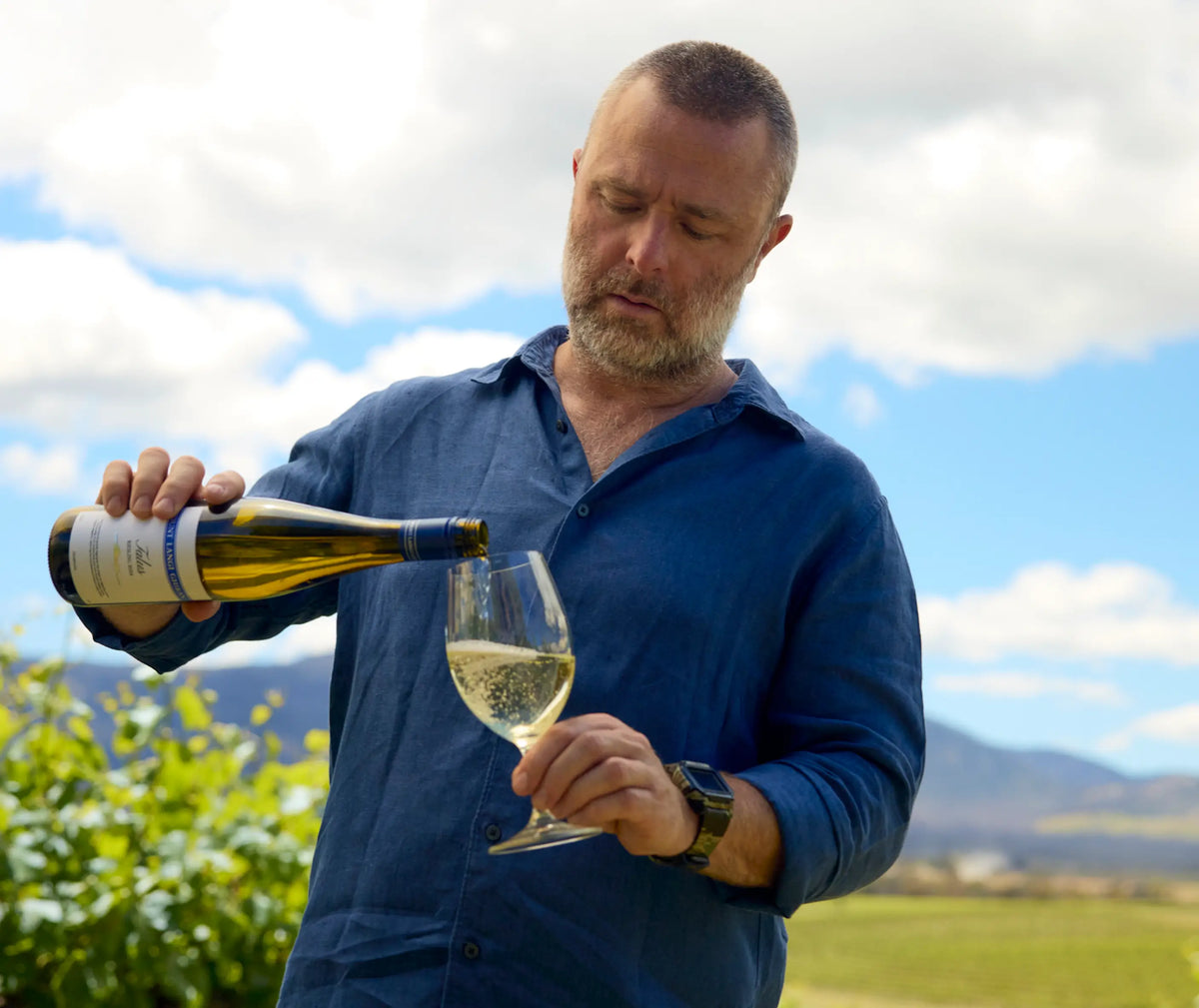 Adam, a Mount Langi Ghiran team member, pouring a glass of white wine outdoors with vineyard hills behind him