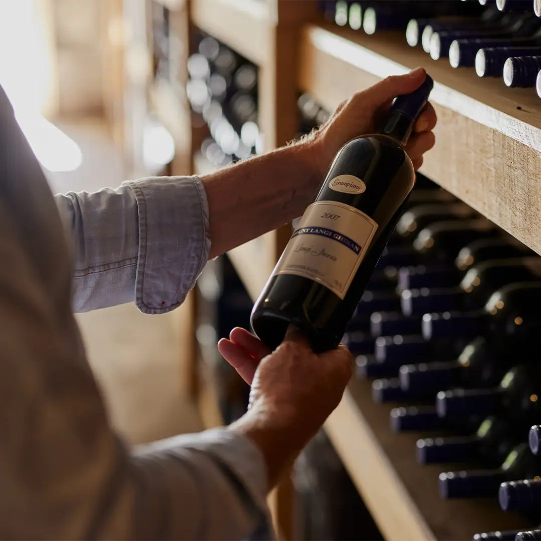 Hands holding a bottle of Mount Langi Ghiran wine on a cellar shelf lined with bottles