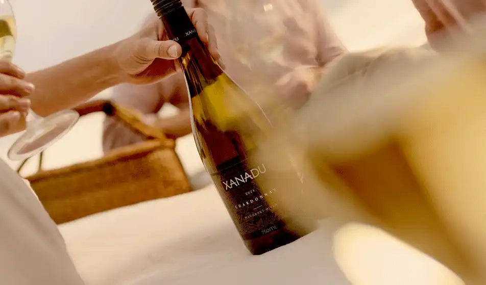 Hands holding a bottle of Xanadu Chardonnay and glasses of white wine at an outdoor picnic
